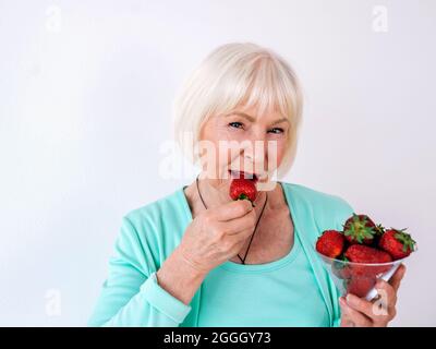 portrait of senior (old) stylish cheerful woman in  turquoise clothes eating strawberries. Summer, travel, anti age, joy, retirement, strawberries Stock Photo