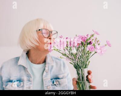 senior stylish woman in glasses and jeans jacket smell carnation flowers in vase. Flowers, retirement, profession, hobby concept Stock Photo