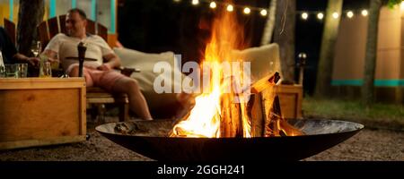 Cast iron fire pit campfire place at forest beach camping with brgiht burning flame at evening time against light bulb garland and trees. Friends sit Stock Photo
