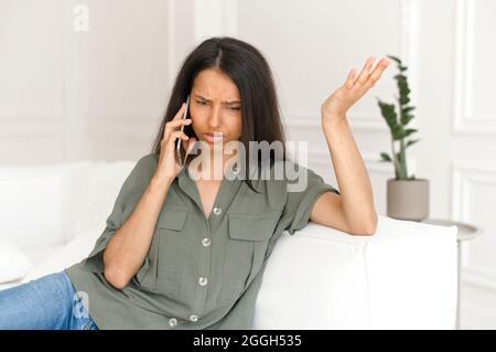 Headshot of confused and annoyed indian woman talking on smartphone sitting on the couch at home, arguing with support services, upset mixed-race lady has unpleasant phone conversation Stock Photo