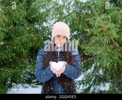 beautiful young woman holding a heart-shaped snowball in her hands, on a snowy winter day in the forest, long red hair, in a hat and gloves Stock Photo