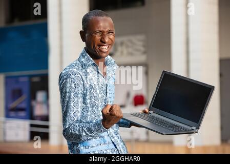 handsome happy african adult man wearing a blue shirt using a computer making a winner gesture in on the street Stock Photo