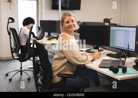 Portrait of smiling caucasian female creative at work, sitting at desk, looking to camera Stock Photo
