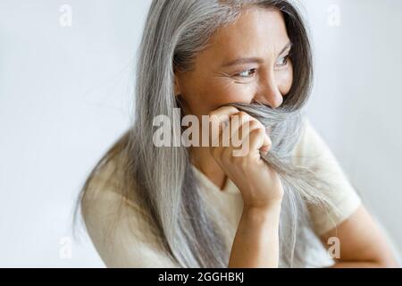 Middle aged Asian woman hides face with grey hair sitting on light background Stock Photo