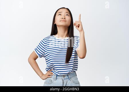 Unsure young asian woman pointing and looking up with troubled, skeptical face expression, having doubts about product or company advertisement Stock Photo