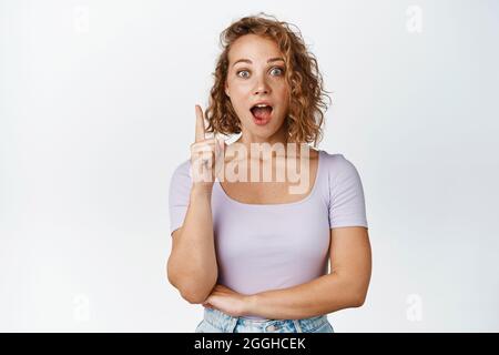 Surprised blond girl with curly hair making announcement, looking amazed, pointing finger up at advertisement, standing in tshirt over white Stock Photo