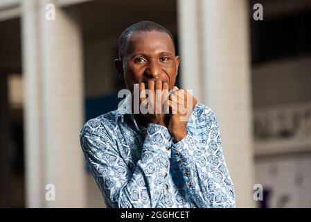 African stressed and nervous adult man with hands on mouth while biting his nails in the street Stock Photo