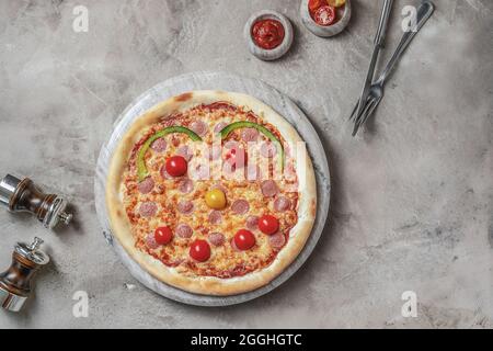 Tasty pizza with  sausage, mozzarella for children with smiley  on marble round tray. Garnished on grey stone backdrop with pizza ingredients Stock Photo