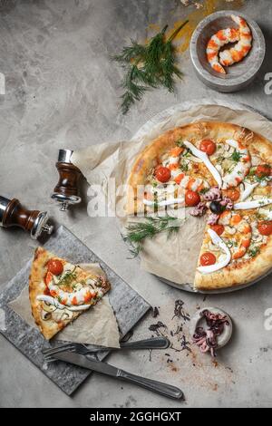 Tasty pizza with  tuna, seafood, shrimps and mini octopus on marble round tray. Garnished on grey stone backdrop with pizza ingredients Stock Photo