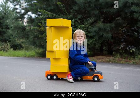 cute smiling boy 5 years old in a blue overalls sits on a large toy car - a truck with many yellow cardboard boxes. Parcel delivery, postman, little t Stock Photo