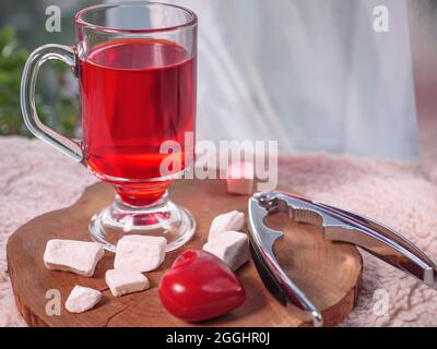 A glass of red berry tea and hazelnuts with nutcracker lying on a wooden board on a cozy orange warm cloth. Window with curtain lace at the background Stock Photo
