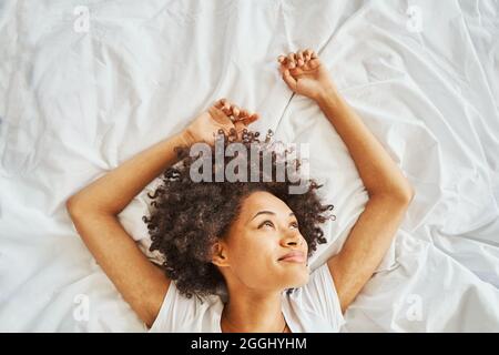 Smiling contented dreamy attractive young African American woman lying on her back on the crumpled bedsheet Stock Photo