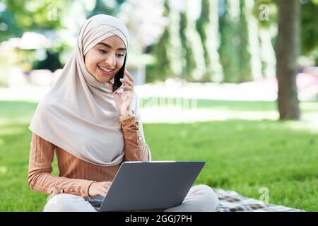 Glad attractive young middle eastern female in hijab looks in laptop, call by phone, works remotely in park Stock Photo