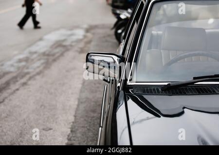 Front-side view of classic black car parked along a street. Close-up vintage chrome car with wing mirror. People walking on the street. Stock Photo