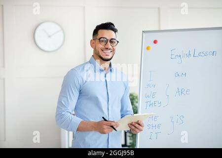 Arab male English teacher explaining rules near blackboard, standing with clipboard, smiling at camera in office Stock Photo