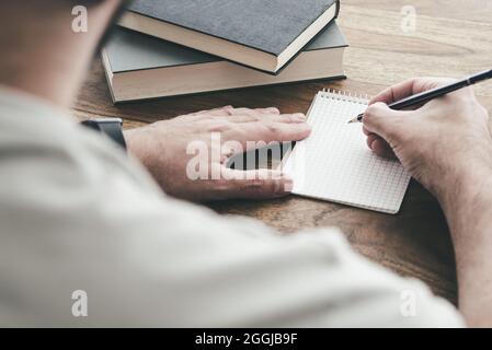 over the shoulder view of man writing on small spiral notepad at wooden table Stock Photo