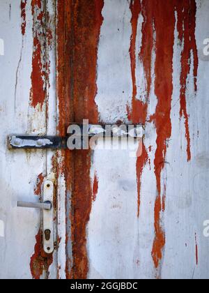 Rust-streaked old white door with metal latch and handle, showing peeling paint and weathered industrial texture. Stock Photo