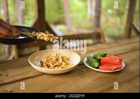 Hands close-up put fried potatoes from frying pan in a plate on wooden table. Fresh farmer eco cucumbers and tomatoes from the vegetable garden. Dinner at countryside yard outdoors Stock Photo