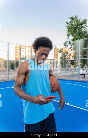 Young afro american basketball player using phone while holding the ball under his arm in the street basketball court. Stock Photo