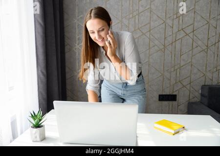 Smiling young female freelancer, employee is standing near workplace, talking on the phone and check some information on the laptop screen. Busy businesswoman, secretary in smart casual outfit indoor Stock Photo