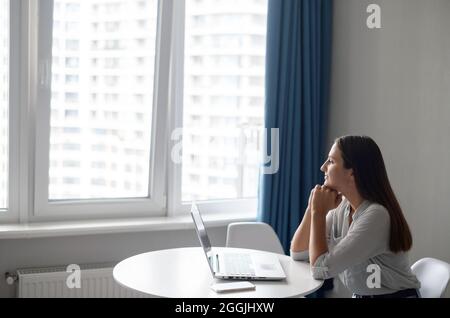Carefree and tranquil young woman sitting at the desk and using laptop computer looks away dreamily, female freelancer looking for inspiration Stock Photo