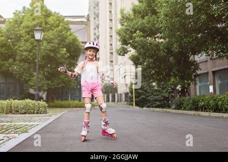 Athletic caucasian girl in pink helmet and knee pads learns to roller skate outdoor at hot summer day. Children education during summer vacation Stock Photo