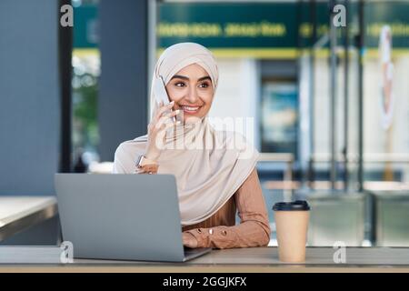 Busy smiling attractive young arab lady in hijab calls on phone, sits with laptop and coffee in cafe Stock Photo