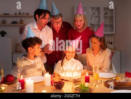 Joyful little girl receiving tasty birthday cake with candles from loving family, celebrating b-day at home in evening Stock Photo