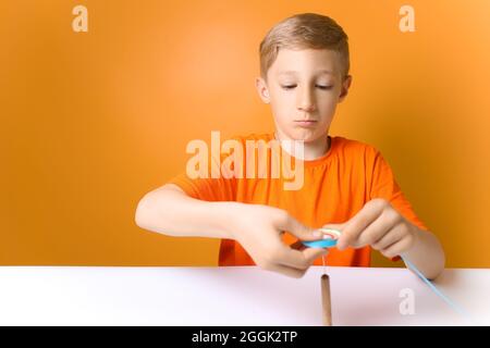 a child in an orange T-shirt sits at a table and twists thin paper strips on a quilling tool Stock Photo