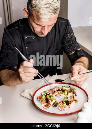 A stylish chef in a black jacket prepares tasty salad with ripe figs, goat Chevre cheese, fresh cucumber roll, beetroot and mustard leaves with tomato Stock Photo