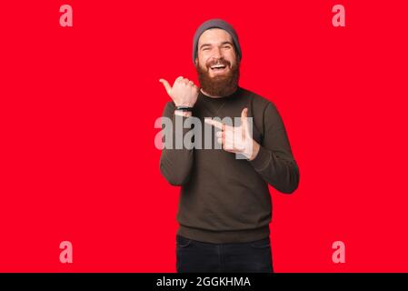 Bearded man wearing warm hat and sweater is pointing at his wrist watch over red background. Stock Photo