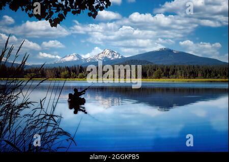 Float tube fly fisherman on Black Butte pond with Three Sisters Mountains. Oregon. Stock Photo