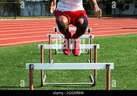Front view of an African American high school boy jumping over track hurdles on a turf field during strength and agility practice. Stock Photo