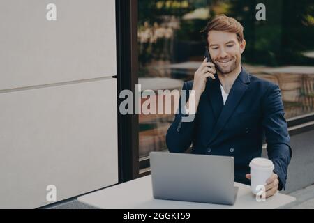 Young successful business owner working on laptop in cafe outside and talking on mobile phone Stock Photo