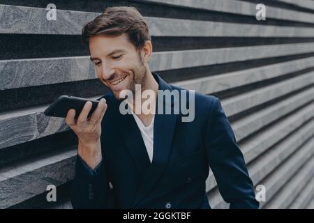 Young attractive man stands outside and using voice recognition assistant on smartphone Stock Photo