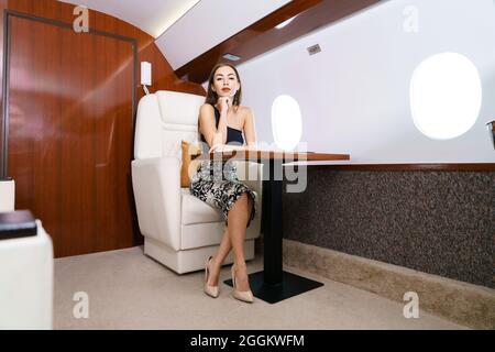 Ideal woman. A modern woman in stylish clothes is smiling while sitting by the window in a first class plane. Beautiful caucasian girl Stock Photo