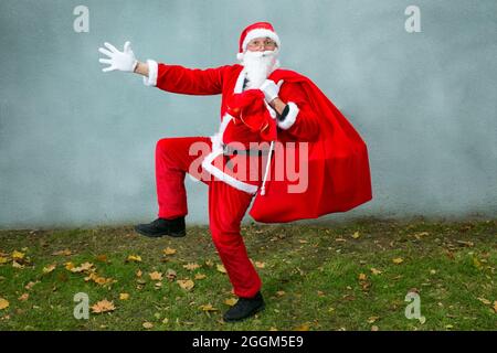 Santa Claus runs with a big red bag with gifts. Close-up on a gray background and green grass. Stock Photo