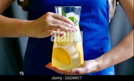 Lemon lemonade with mint, lemon and ice in a carafe. Woman in a blue apron holds a cooling, refreshing, fruity, summer drink. Stock Photo