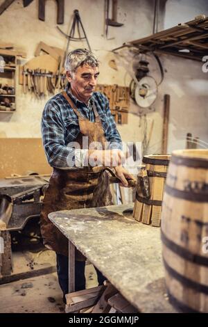 Carpenter places metal stripe on a new wooden barrel made in his cozy workshop. Stock Photo