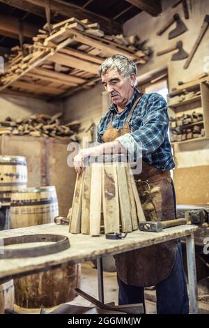 Old craftsman builds a new wooden barrel from metal and lumber in an old-fashioned way. Stock Photo
