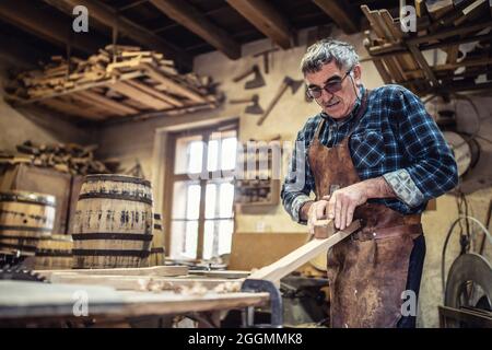 Older man smothes the surface of lumber before using it to build wooden barrels. Stock Photo