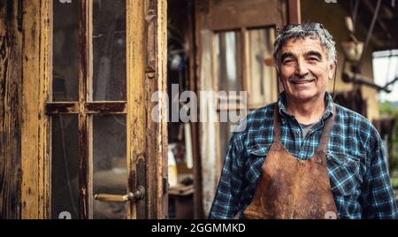 Aged craftsman standing and smiling in front of his rustic workshop. Stock Photo