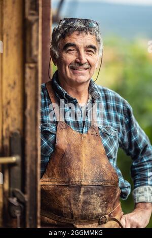 Older and experienced craftsman stands smiling in front of the workshop in checkered shirt and leather apron. Stock Photo