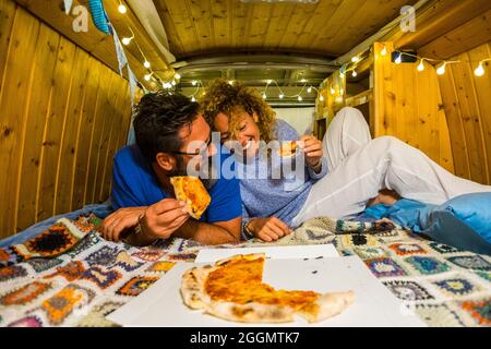 Happy couple have fun eating pizza inside an hand made van camper vehicle - travel and vacation lifestyle caucasian people laugh a lot Stock Photo