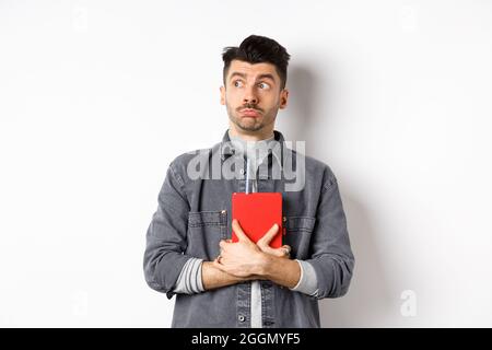 Silly young man looking aside at logo with timid face, holding his diary pressed to chest, carry journal, standing on white background Stock Photo