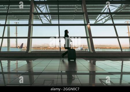 Silhouette of female flight passenger with luggage walking at airport terminal. Woman traveler walking in front of large window at airport terminal. Stock Photo