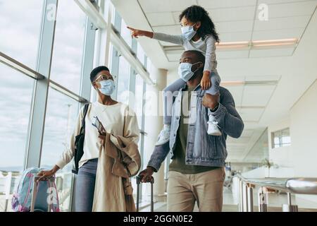 Young girl pointing away while on shoulder of her father and woman at airport. African family traveling during pandemic. Stock Photo