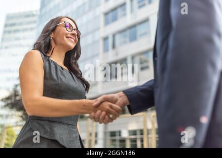 Two people in business clothes shake hands surrounded by office buildings. Stock Photo