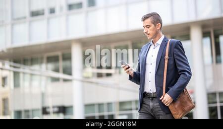 Man in casual business attire checks his phone walking outdoors in an office buildings environment. Stock Photo
