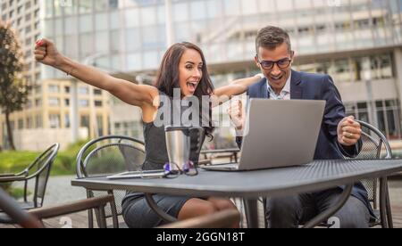 Male and female coworkers showing thrilled enthusiasm over successful deal just done on a laptop in fron of them outdoors in the city. Stock Photo